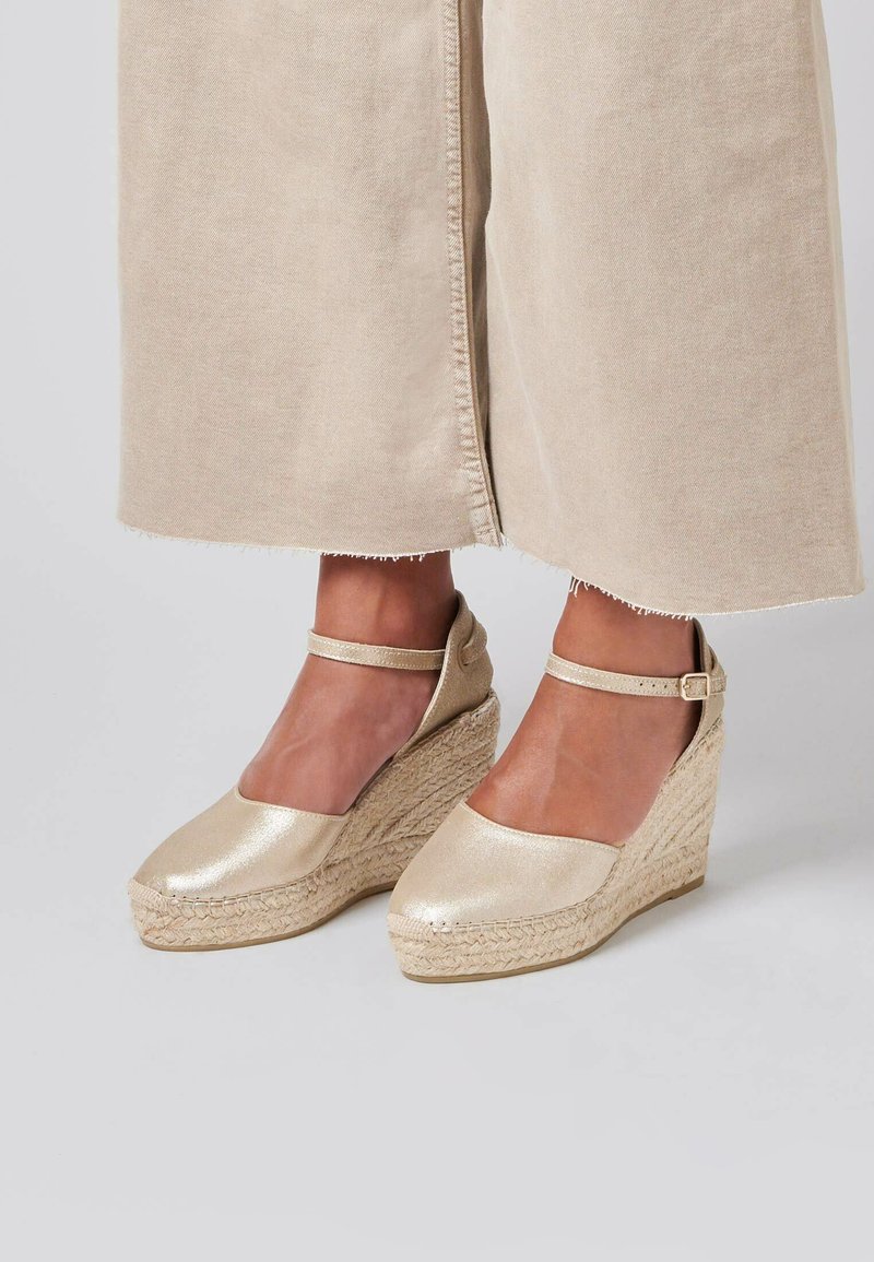 Des chaussures compensées dorées en espadrille avec une bride à la cheville, présentant une semelle compensée en jute texturé et un dessus en métal lisse.