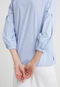 Blouse rayée bleu clair avec des manches bouffantes, des accents de boutons et un ourlet légèrement incurvé. Associée à un pantalon blanc, les mains croisées dans le dos.