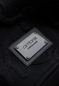 Tessuto nero con un motivo testurizzato, con un'etichetta rettangolare in pelle nera impreziosita dalla scritta "ombre menswear," fissata con quattro rivetti metallici.