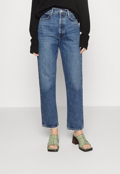 Jeans z wysokim stanem w niebieskim kolorze, prosty krój, postrzępione wykończenie, w połączeniu z zielonymi sandałami na obcasie z krzyżującymi się paskami i brązowym obcasem.