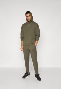 Olivgrüner Kapuzenpullover mit Zugband, mit Bündchen an den Ärmeln und Logo auf der Vorderseite, kombiniert mit passender Jogginghose. Schwarze Sportschuhe runden das Outfit ab.