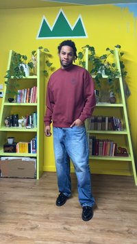 Hanorac bordo cu manșete striate, blugi largi de culoare albastru deschis și pantofi negri. Fundalul prezintă rafturi verzi și un perete galben.