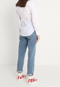 Biała koszula z długim rękawem i guzikami, niebieskie dżinsy z wysokim stanem oraz białe sneakersy z czerwonym tekstem. Gładki materiał, luźny krój, podwinięte mankiety.