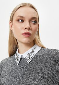 Top in bianco e nero con texture, colletto bianco decorato con embellishments argento e nero. Presenta un collo rotondo e maniche lunghe.