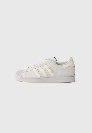 Zemu Adidas Superstar ar zemu apakšējo daļu gaismas bēšā krāsā ar krēmkrāsas svītrām, gumijas čības purngala daļu un zelta burtiem "Superstar" sānos.