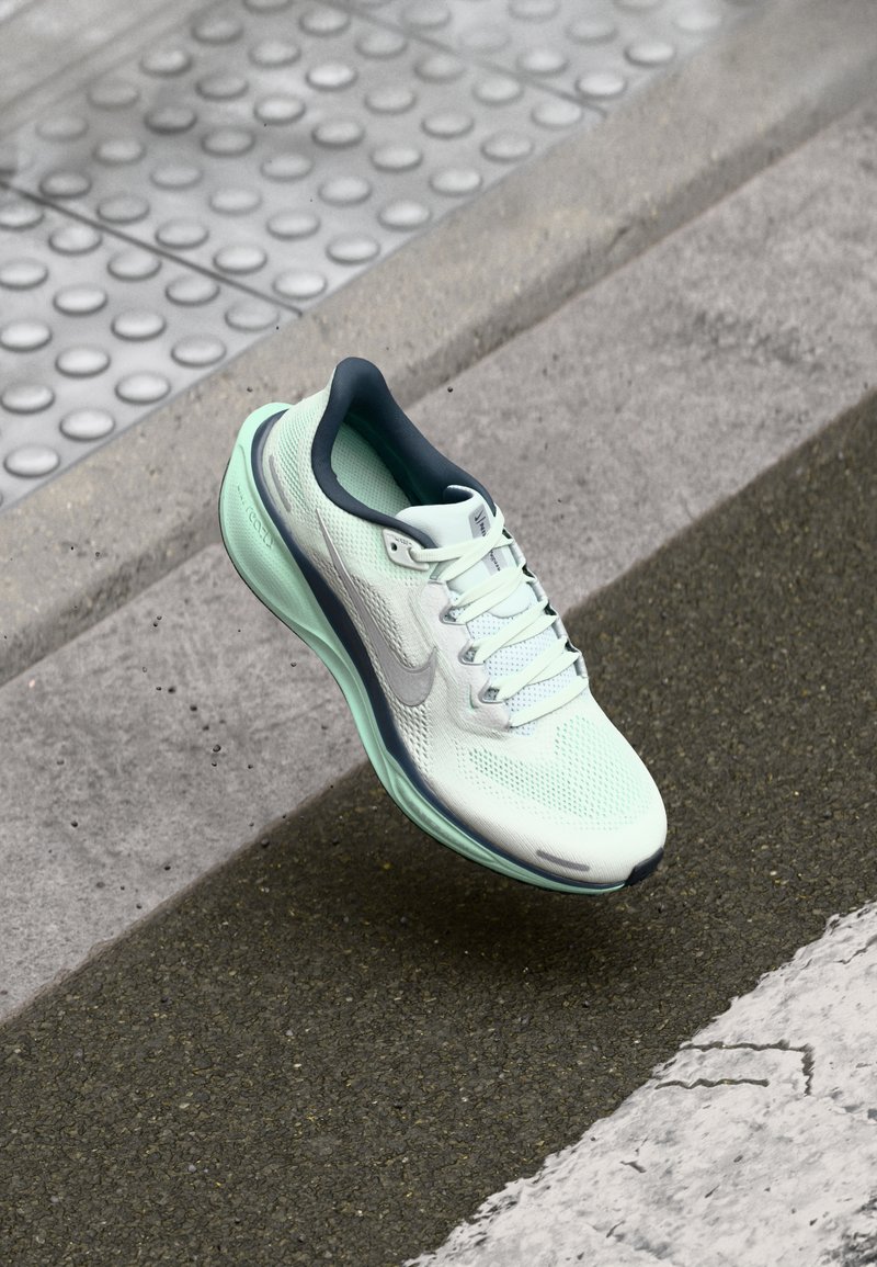 Zapatilla deportiva de color verde claro con una parte superior de malla transpirable, suela texturizada y acentos reflectantes. Cuenta con un interior y cordones de color azul oscuro.