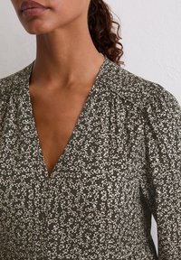 Ciemnozielona sukienka w kwiaty z dekoltem w kształcie litery V, bufiastymi rękawami i teksturowanym materiałem. Posiada białe wzory kwiatowe na całej powierzchni.