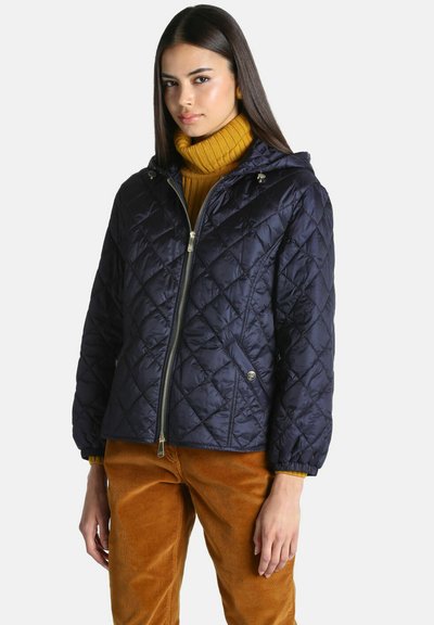Femme portant une veste matelassée bleu marine avec capuche par-dessus un pull à col roulé jaune et un pantalon en velours marron, debout devant un fond uni.