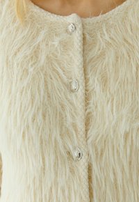 Close-up van een crèmekleurige, fuzzy gebreide vest met zilveren decoratieve knopen op de bovenste helft van een persoon.