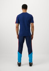 Jūras spēku sporta krekls un bikses ar sarkaniem un dzelteniem sānu svītrām. Biksēm ir zilā apakšējā daļa. Audums izskatās gluds un pieguļošs.