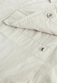 La tela de color gris claro presenta una textura arrugada, con un borde doblado que muestra un hardware de presión plateado y detalles de costura en contraste.