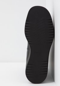 Suela de goma negra con un patrón texturizado, con un material superior gris y suave. Marca visible en la suela.