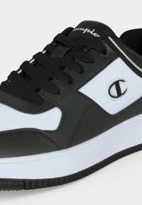 Baskets basses avec panneaux noirs et blancs, lacets noirs, logo brodé « C » sur le côté, et semelle blanche avec semelle extérieure noire.