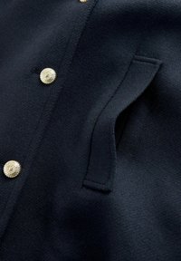 Haditengerészeti gyapjú kabát aranyszínű gombokkal, texturált felülettel és egy oldalsó zsebbel, amely sima varrási részletet tartalmaz.