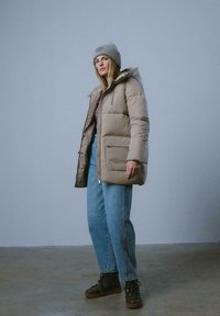 Kurtka puchowa w jasnym beżu z kapturem, z zamkiem błyskawicznym i dwiema przednimi kieszeniami. Połączona z luźnymi niebieskimi dżinsami i ciemnymi sznurowanymi butami.