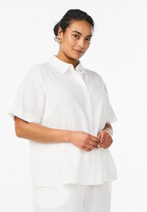 Naine kannab lahtist valget lühikeste varrukatega nööpidega särki ja sobivaid valgeid pükse, poseerides käed ees kokkusirutatud.