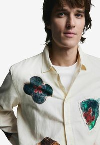 Valge nööbitav särk, millel on värvilised kangast plaastrid, mis kujutavad lillemustreid rohelises, punases ja pruunis toonis. Pehme, tekstuuriga puuvillane materjal.