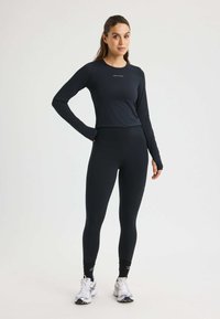 Black long-sleeve fitted top with logo, paired with high-waisted black leggings. Model stands in athletic shoes, showcasing a sleek, fitted design.