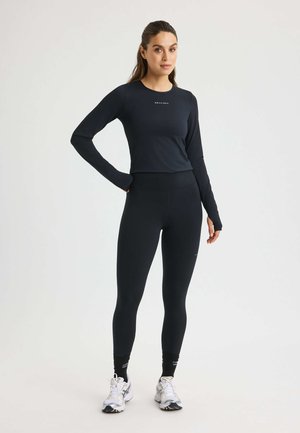 Zwarte, langesleeve, getailleerde top met logo, gecombineerd met hooggerokte zwarte leggings. Het model staat in sportieve schoenen en toont een slank, getailleerd ontwerp.