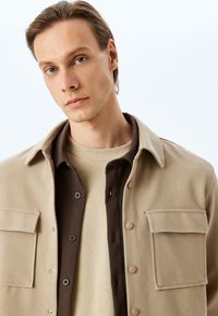 Beige button-up jakke med to forreste lommer og brune detaljer over en lys sweater. Glat tekstur og enkelt design.