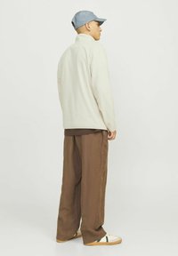 Beige fleece zip-up jacka, avslappnad passform, tillsammans med lösa bruna byxor och vita sneakers med gröna accenter. En ljusblå keps fulländar looken.