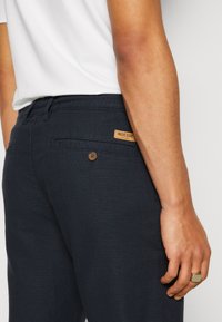 Dark blue pants with a textured fabric, featuring a back pocket with a brown button and a tan logo patch near the waistband.
