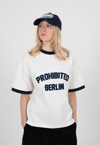 Femme aux cheveux blonds portant un t-shirt blanc "Prohibited Berlin" et une casquette bleu marine, regardant sur le côté avec les mains dans les poches.