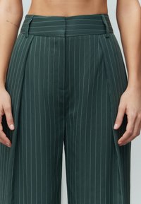Groene gestreepte broeken met hoge taille, voorzien van plooien, een soepele textuur en zijzakken. Geaccentueerd met subtiele blauwe lijnen.