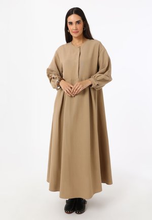 Frau mit langen dunklen Haaren trägt ein lockeres beiges langes Kleid und schwarze Clogs, steht mit verschränkten Händen in der Taille vor weißem Hintergrund.