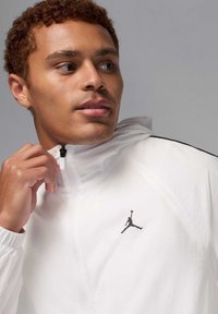 Chaqueta cortavientos blanca con detalles en negro y un pequeño logo Jumpman. Cuenta con un cuello con cremallera y tela ligera y texturizada.