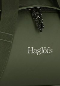 Borsone verde impermeabile con superficie texturizzata e chiusura a coulisse nera, con il logo ricamato "Haglöfs" in bianco.