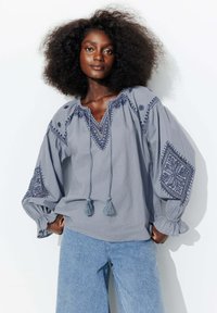 Chemise brodée gris clair avec des manches bouffantes, présentant des liens à pompons et des motifs complexes sur les manches. Associée à un jean bleu clair.