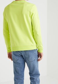 Felpa a maniche lunghe, di un brillante verde neon, con un collo rotondo, texture liscia e orlo aderente, abbinata a jeans azzurri chiari.