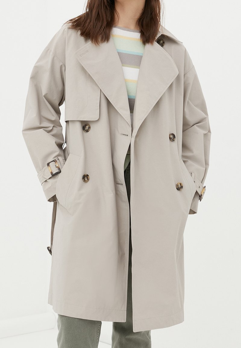 Femme portant un trench-coat beige croisé avec de larges revers, des poches latérales, des poignets boutonnés, par-dessus un pull rayé et un pantalon vert.