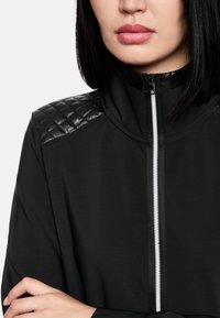 Schwarze Jacke mit hohem Kragen, ausgestattet mit gesteppten, glänzenden Akzenten auf den Schultern und einem weißen Reißverschluss auf der Vorderseite. Glatte Textur des Stoffes.