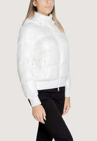 Giacca a vento bianca con cappuccio, chiusura frontale con zip e polsini a costine. Il materiale esterno ha una texture lucida e presenta una tasca laterale con zip.