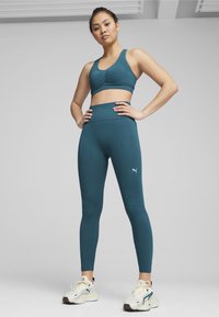 Teal športni modrček s sprednjim nabiranjem, usklajen s teal visokimi pajkicami. Obe sta izdelani iz raztegljive tkanine; bel logo Puma na strani.