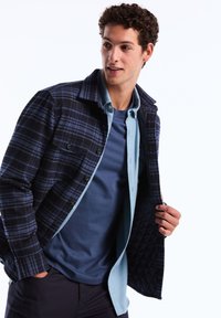 Marineblaues und schwarzes karierter Jacket über einem hellblauen Hemd und einem dunkelblauen T-Shirt; sichtbare Knopfdetails, gefüttertes Innenfutter und strukturierter Kragen.