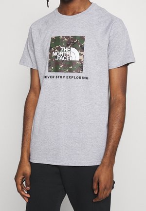 Camiseta gris de manga corta con un logo cuadrado de camuflaje y texto en negro que dice "NUNCA DEJES DE EXPLORAR", hecha de tela de algodón.