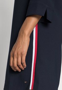 Marineblaues Kleid mit entspannter Passform, ausgestattet mit roten und weißen Streifen an der Seite. Zusätzliche Details umfassen ein kleines Markenlogo am Saum.