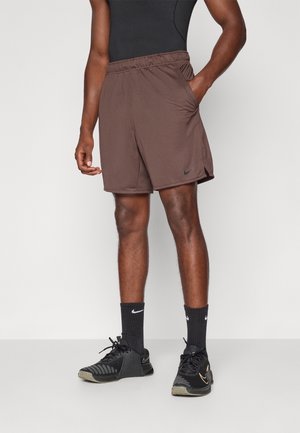 Mand iført brune atletikshorts med lommer, sort shirt, sorte Nike-strømper og sorte Nike-sneakers, der står mod en ensfarvet baggrund.