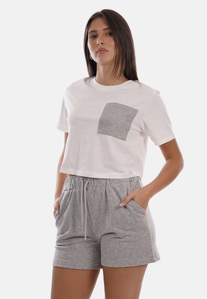 Donna che indossa una t-shirt corta bianca con taschino grigio sul petto e pantaloncini grigi con coulisse coordinati, in piedi con le mani nelle tasche.