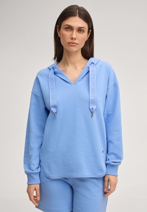 Młoda kobieta w jasnoniebieskiej bluzie z szerokimi sznurkami i pasujących szortach, stojąca na neutralnym tle.