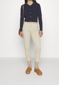 Suéter recortado azul marino con cuello y botones grandes, combinado con pantalones ajustados de color beige claro y zuecos marrones con clavos decorativos.