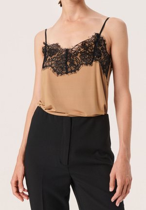 Kobieta ubrana w beżowy top na cienkich ramiączkach z czarną koronką, w zestawie z czarnymi spodniami z wysokim stanem, stojąca na tle jednolitego koloru.