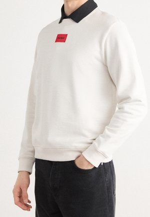 Mies pukeutunut off-white-väriseen pitkähihnaiseen collegepaitaan, jossa on musta kaulus ja punainen suorakulmainen logopläkki, yhdistettynä mustiin housuihin, käsi taskussa.