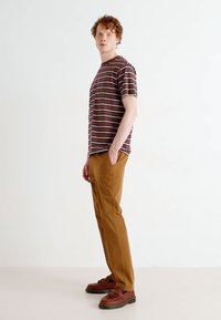 Camisa de manga corta a rayas en marrón y blanco, combinada con pantalones color beige y zapatos marrones, exhibida en un modelo frente a un fondo neutro.