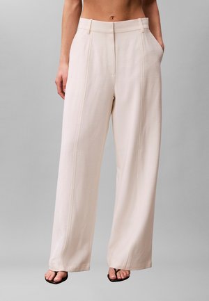 Femme portant un pantalon crème taille haute, jambes larges avec un détail de couture sur le devant et des sandales noires à bout carré, mains dans les poches.