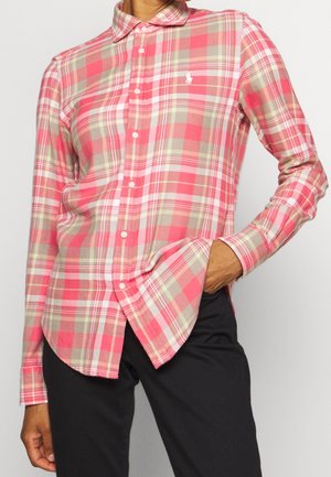 Mężczyzna w różowo-beżowej kraciastej koszuli na długi rękaw z małym białym logo na piersi, w zestawieniu z czarnymi spodniami. Ręka w kieszeni.