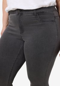 Primer plano de una persona que lleva jeans ajustados de color gris oscuro y una camisa blanca metida por dentro, centrado en la cintura y la parte superior del muslo.